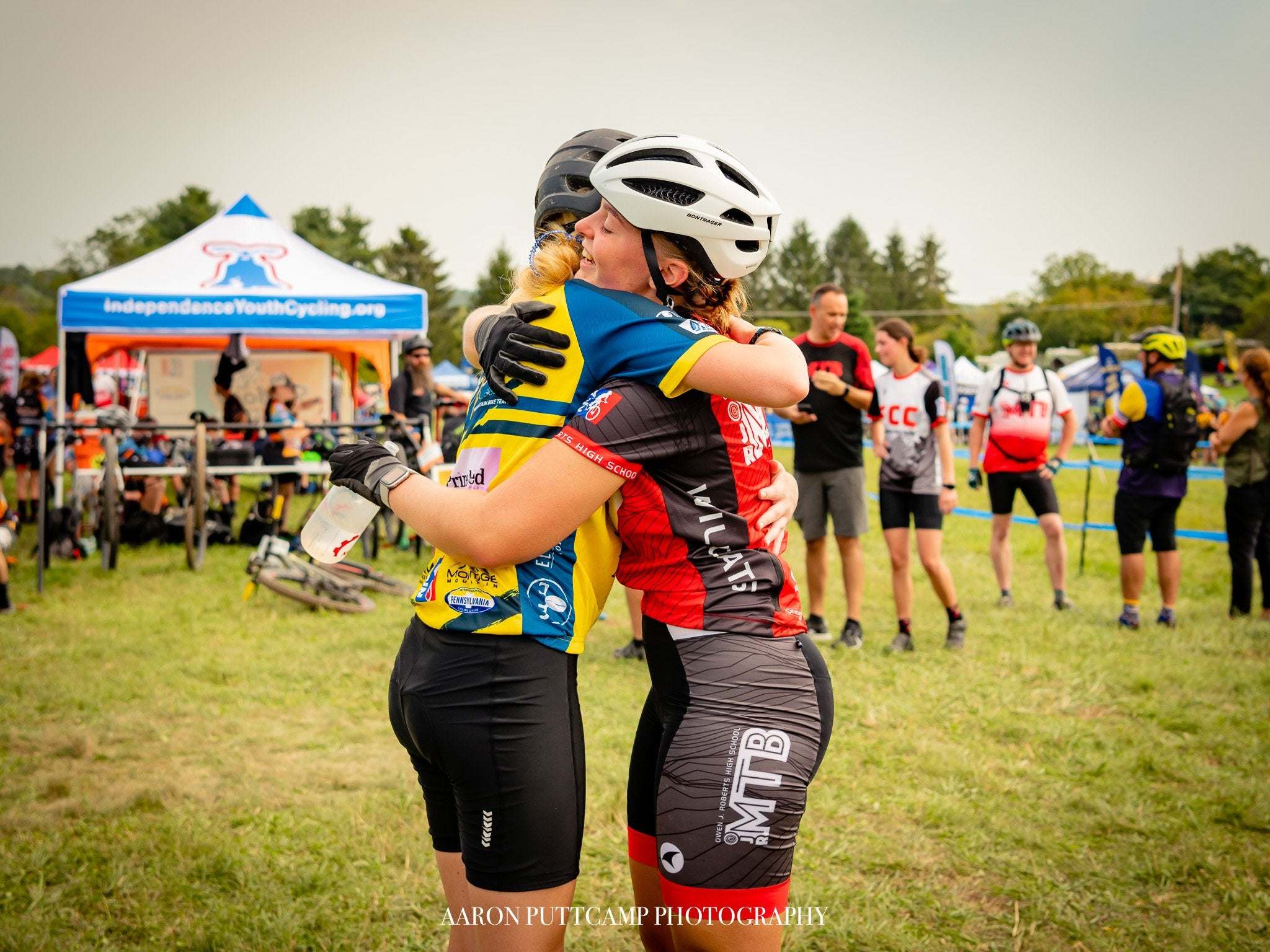 Two cyclists wearing helmets hugging at a youth cycling event supporting PICL with people and tents in the background